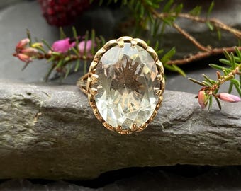 Pode incluir: Um anel de ouro com uma grande gema oval transparente engastada em uma delicada moldura ornamentada.