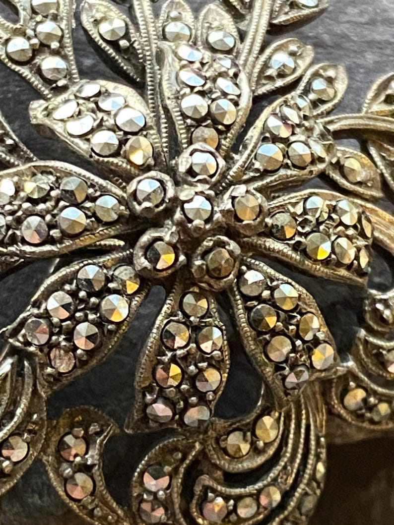May include: Close-up of a silver floral brooch. The brooch has a detailed design with layers of stylized leaves and flower petals, each with small, sparkling, round stones. The metal has an antique finish.