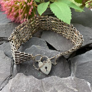 Könnte beinhalten: Silberfarbenes Armband mit gewebtem Design und herzförmigem Vorhängeschloss. Das Armband besteht aus miteinander verbundenen rechteckigen Gliedern, die ein flexibles, strukturiertes Aussehen erzeugen. Eine kleine Kette verbindet das Vorhängeschloss mit dem Armband.