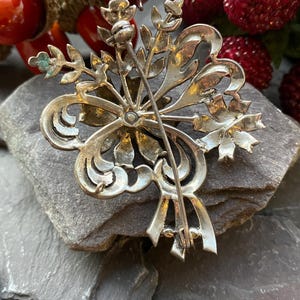 May include: A silver brooch with an elaborate floral design. The brooch showcases stylized leaves, scrolls, and a central pin. The reverse side of the brooch is visible, revealing the pin mechanism and a textured surface.