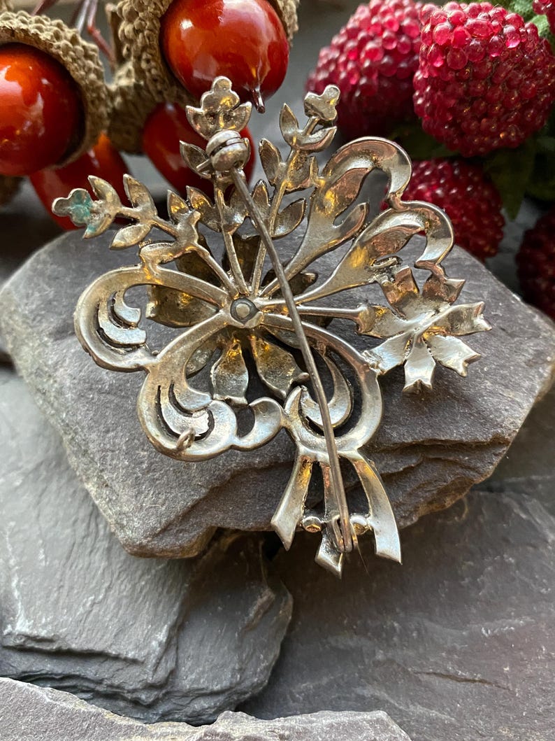 May include: A silver brooch with an elaborate floral design. The brooch showcases stylized leaves, scrolls, and a central pin. The reverse side of the brooch is visible, revealing the pin mechanism and a textured surface.