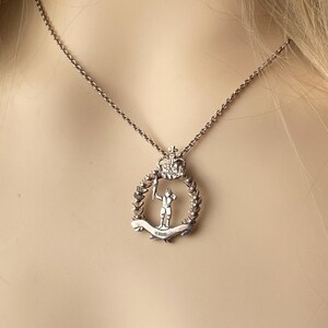 May include: A silver-toned pendant necklace featuring a circular emblem with a crown at the top. Inside the circle is a figure holding a staff, surrounded by a laurel wreath. The necklace chain is delicate and silver.