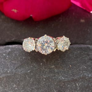 Può includere: Un anello a tre pietre con tre grandi gemme rotonde e chiare. L'anello ha una fascia d'oro. Le gemme brillano. L'anello è esposto su una superficie scura e strutturata.