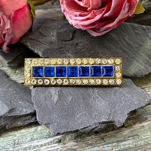 May include: A rectangular gold-tone brooch featuring a central row of seven square-cut deep blue stones. The blue stones are bordered by two horizontal rows of small, round, clear white rhinestones set into the metal frame of the jewelry piece.