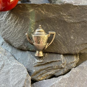 May include: A miniature silver teapot with detailed floral patterns and a ribbed lower section. The teapot features a handle, spout, and a domed lid with a knob. It is displayed on a grey stone surface. The teapot is a charming decorative item.