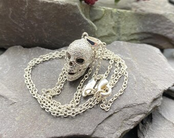 Puede incluir: Un collar de cadena de plata con un colgante de calavera de plata incrustado con piedras de circonita cúbica transparentes. La calavera tiene ojos de esmalte negro.
