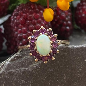 May include: A gold ring with a white opal gemstone surrounded by a halo of small purple gemstones.