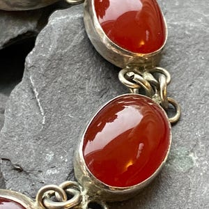 Pode incluir: Uma pulseira de prata com pedras preciosas ovais, polidas, vermelho-alaranjadas. As pedras s&atilde;o engastadas em uma moldura de prata e unidas por elos de corrente de prata. A pulseira &eacute; exibida em uma superf&iacute;cie cinza texturizada.