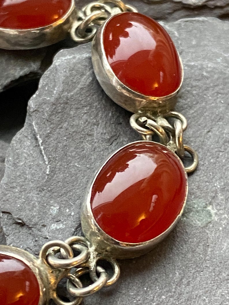 Pode incluir: Uma pulseira de prata com pedras preciosas ovais, polidas, vermelho-alaranjadas. As pedras s&atilde;o engastadas em uma moldura de prata e unidas por elos de corrente de prata. A pulseira &eacute; exibida em uma superf&iacute;cie cinza texturizada.