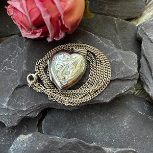 Könnte beinhalten: Ein silberfarbener herzförmiger Medaillon-Anhänger an einer zarten passenden Kette. Das Medaillon weist auf der Vorderseite ein kompliziertes geprägtes florales Rankenmuster und am Ende der Kette einen kleinen runden Verschluss auf.