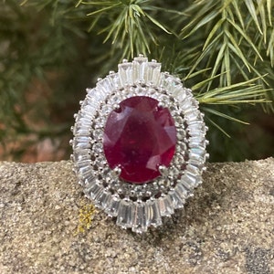 May include: A silver ring with a large oval-shaped ruby gemstone surrounded by smaller clear gemstones.