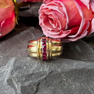 May include: A gold ring featuring a central vertical row of four square-cut deep red rubies. The band has a wide, multi-layered design with polished gold surfaces that frame the channel-set gemstones in the center of the piece.