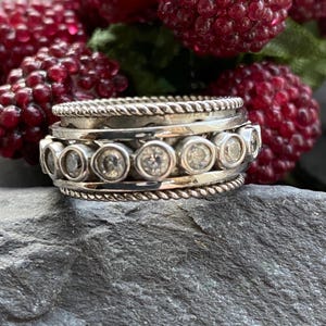 May include: A silver ring with a textured design and clear gemstones. The ring has a braided rope-like border and a row of circular gemstones. The ring is displayed against a dark background with red berries.