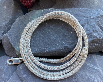 May include: A silver chain necklace with a woven design. The chain is coiled on a gray stone surface.