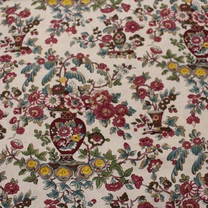 Op de afbeelding: Een crèmekleurige stof met een herhalend patroon van donkerrode urnen gevuld met levendige bloemstukken. Het ontwerp bevat dieprode, roze en gele bloesems, groene bladeren en blauwgroene accenten voor een dicht botanisch motief.