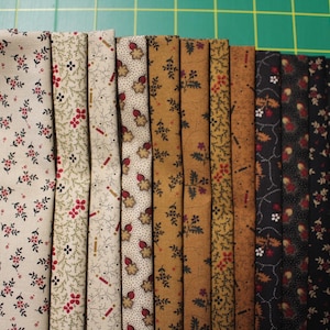 May include: A collection of twelve folded fabric strips arranged in a gradient from light cream to dark black. Each strip features a distinct floral or geometric pattern in shades of red, brown, tan, and black, showcasing a variety of small-scale prints.