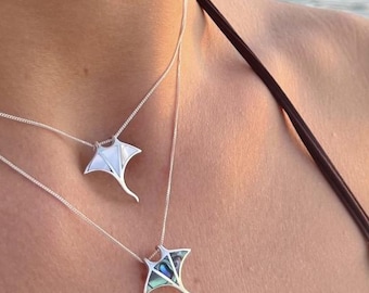 May include: Two silver necklaces with stingray pendants. The pendants have a white and iridescent blue-green design. The necklaces are worn against a person's skin, with a blurred ocean background.