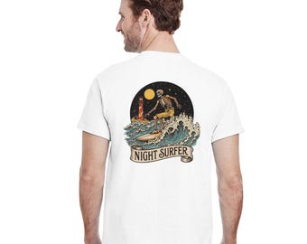 May include: White t-shirt featuring a graphic of a skeleton surfing at night. The design includes a lighthouse, moon, and the words "NIGHT SURFER" on a banner. The surfer is wearing yellow shorts.