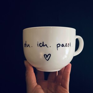 Könnte beinhalten: Weiße Keramiktasse mit schwarzem handschriftlichem Text "du. ich. passt." und einem kleinen Herz.