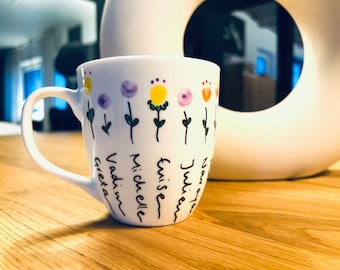 May include: White ceramic mug with a black outline of flowers and the names Greta, Vadim, Michelle, Twise, Julien, and Nora written on the side.
