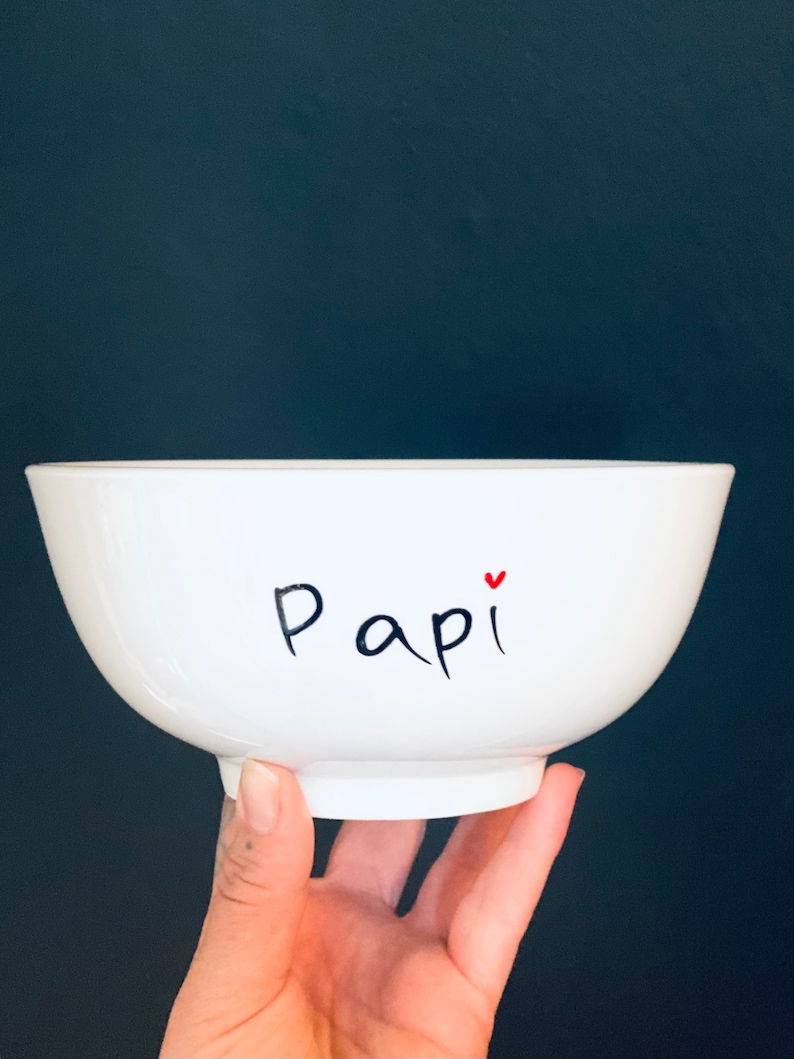 May include: A white ceramic bowl with the word "Papi" written in black script, accented with a small red heart. The bowl has a rounded shape and a small base. The bowl is held in front of a dark blue background.