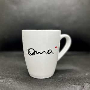 Könnte beinhalten: Weiße Keramiktasse mit einer schwarzen, kursiv geschriebenen Aufschrift "Oma" mit einem kleinen roten Herz.