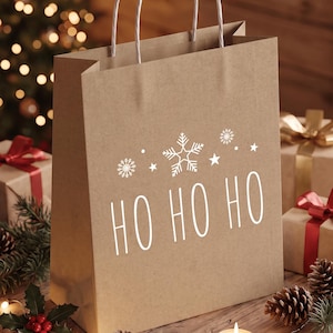 Könnte beinhalten: Eine braune Geschenktüte aus Kraftpapier mit weißen gedrehten Papiergriffen. Auf der Vorderseite befindet sich ein weißer Grafikdruck mit Schneeflocken, Sternen und dem Text HO HO HO. Die Tüte ist aufrecht stehend mit einem rechteckigen Boden.