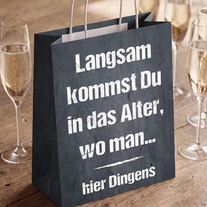 May include: A dark gray paper gift bag with white text that reads: Langsam kommst Du in das Alter, wo man... hier Dingens. The bag features two white twisted paper handles and has a textured surface appearance resembling a chalkboard finish.