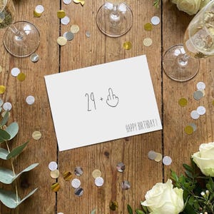 May include: A white rectangular greeting card with black text and a line drawing. The card features the number 29, a plus sign, an illustration of a hand with a raised middle finger, and the text HAPPY BIRTHDAY printed in a simple font.