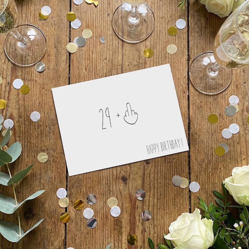 May include: A white rectangular greeting card with black text and a line drawing. The card features the number 29, a plus sign, an illustration of a hand with a raised middle finger, and the text HAPPY BIRTHDAY printed in a simple font.