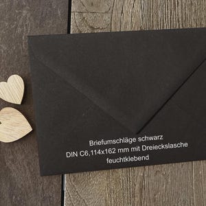 May include: A black envelope with a triangular flap and two small wooden heart cutouts. The envelope features white printed text: Briefumschl&auml;ge schwarz, DIN C6, 114x162 mm mit Dreieckslasche, feuchtklebend. The envelope is rectangular with a pointed flap.