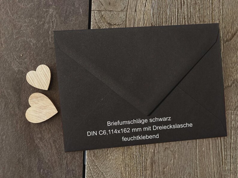 May include: A black envelope with a triangular flap and two small wooden heart cutouts. The envelope features white printed text: Briefumschl&auml;ge schwarz, DIN C6, 114x162 mm mit Dreieckslasche, feuchtklebend. The envelope is rectangular with a pointed flap.