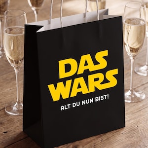 May include: A black paper gift bag with white twisted paper handles. The front features the yellow text "DAS WARS" in a bold, stylized font, with the white text "ALT DU NUN BIST!" printed directly underneath the main yellow lettering.
