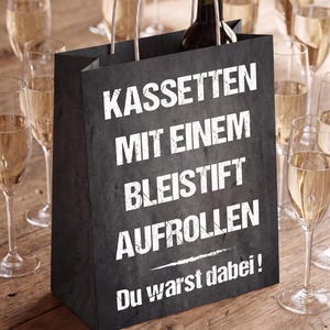 May include: A black paper gift bag with white text that reads KASSETTEN MIT EINEM BLEISTIFT AUFROLLEN Du warst dabei! The bag features white twisted paper handles and contains a dark glass wine bottle with a cork stopper.