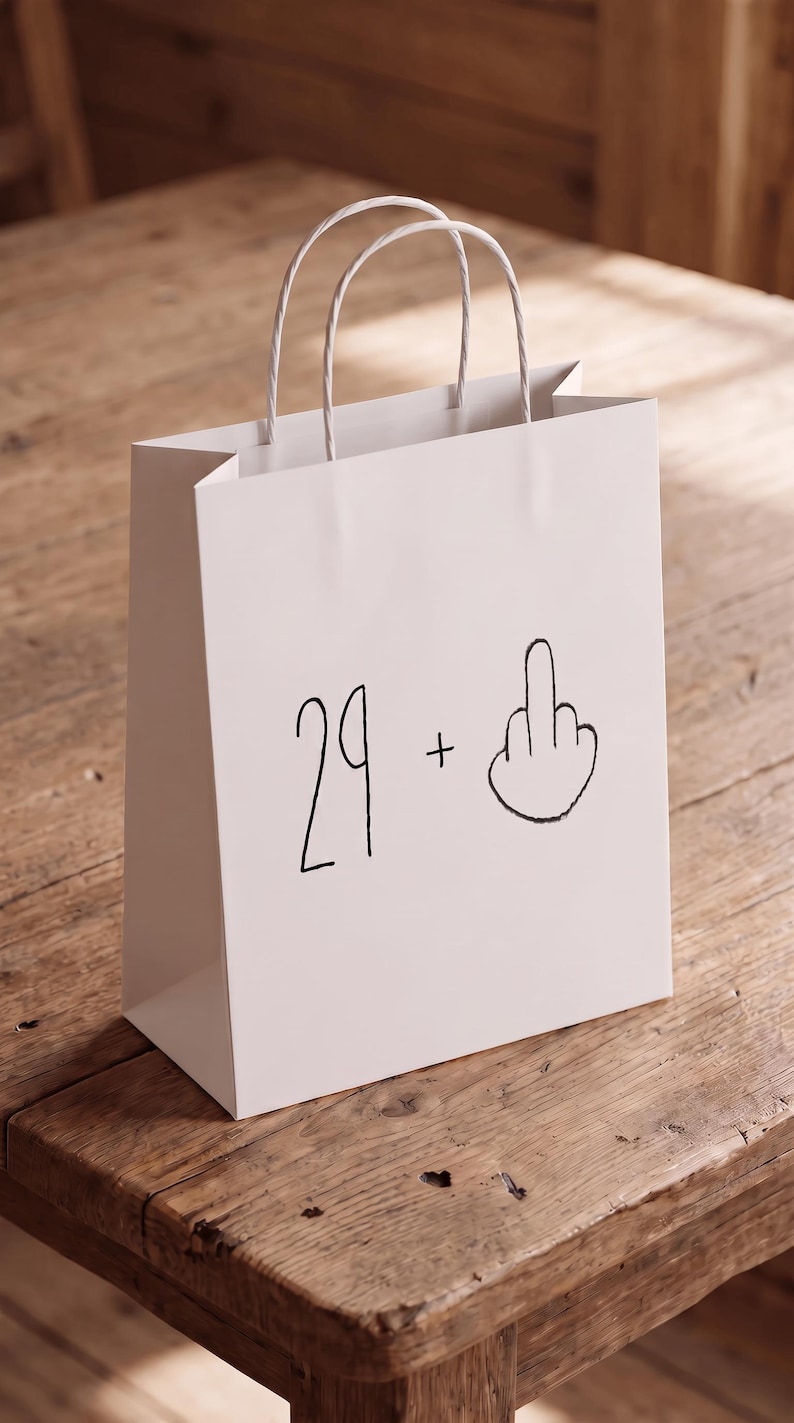 May include: A white paper gift bag with two twisted paper handles. The front of the bag features black printed text that reads 29 + followed by a hand-drawn illustration of a middle finger gesture, creating a humorous graphic design.