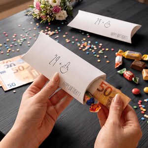 May include: A white rectangular paper gift box with the text 29 + middle finger icon and HAPPY BIRTHDAY printed on the front. The box is being held open to reveal a 50 Euro banknote and a small wrapped candy inside, with confetti scattered nearby.