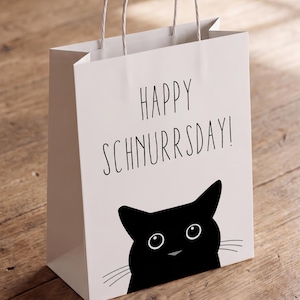 May include: A white paper gift bag with two white twisted paper handles. The front features a black graphic of a cat face and the black text "HAPPY SCHNURRSDAY!" printed in a simple, sans-serif font. The bag has a rectangular base and sides.