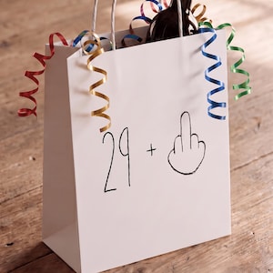 May include: A white gift bag with two white paper handles, featuring the black printed text 29 + and a hand gesture icon. A dark glass bottle with a cork stopper is inside the bag, surrounded by red, gold, blue, and green curling ribbons.