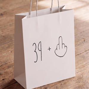 May include: A white paper gift bag with two twisted paper handles. The front of the bag features a black hand-drawn graphic consisting of the number 39, a plus sign, and an illustration of a middle finger gesture centered on the surface.
