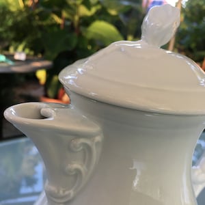 Può includere: Caffettiera in porcellana bianca con coperchio e manico decorativo. Il coperchio ha un piccolo pomo a forma di fiore.