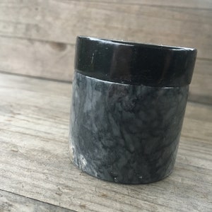 Op de afbeelding: Een kleine, ronde, zwarte en grijze marmeren container met een zwart deksel. De container heeft een marmerpatroon.