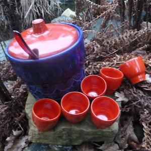 Puede incluir: Un tazón de ponche de cerámica azul y rojo con tapa y cucharón de madera. Seis pequeñas tazas de cerámica roja están dispuestas alrededor del tazón.
