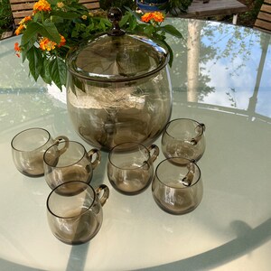 Può includere: Un set di ciotola per punch e tazze in vetro marrone. La ciotola ha un coperchio e le tazze hanno manici. Il set è su un tavolo di vetro.