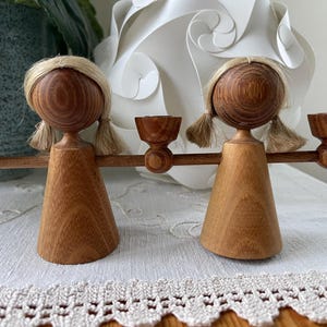 Può includere: Due portacandele a forma di angelo in legno. Ogni angelo ha una testa rotonda con trecce bionde, un corpo conico e braccia tese che tengono le coppe delle candele. Il legno ha un tono caldo e naturale. Su una tovaglia di pizzo bianco.