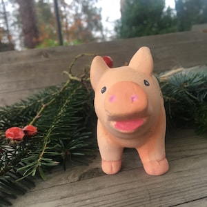 Op de afbeelding: Een keramiek varkensbeeldje met een roze snuit en een roze glimlach. Het beeldje staat op een houten oppervlak met groene dennentakken en rode bessen.