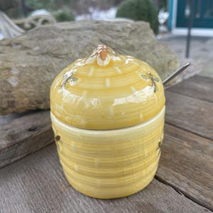 Puede incluir: Tarro de miel de cerámica amarilla con tapa, con ilustraciones de abejas. El tarro tiene una superficie texturizada y se inserta una cuchara pequeña. El tarro de miel está sobre una superficie de madera.