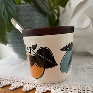 Peut inclure: Pot en céramique crème avec un couvercle en bois foncé et une cuillère en argent. Le pot est décoré d'illustrations de fruits orange, noirs et bleu sarcelle. Un article décoratif pour la cuisine ou la salle à manger.