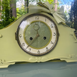 Op de afbeelding: Een vintage, lichtgroene mantelklok met een crèmekleurige wijzerplaat. De wijzerplaat heeft zwarte cijfers en goudkleurige wijzers. Een lichtgroene strik is aan de bovenkant bevestigd. De klok staat op een blauw oppervlak. Ongeveer 25 cm breed.