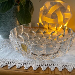 Peut inclure: Un bol en verre transparent à facettes avec un motif géométrique. Le bol repose sur un tissu blanc bordé de dentelle. Une plante en pot et une lumière décorative sont en arrière-plan.
