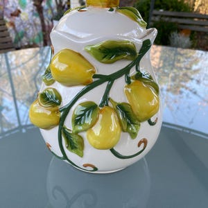 May include: White ceramic jar with a lemon tree design. The jar has a lid and is decorated with green leaves and yellow lemons.